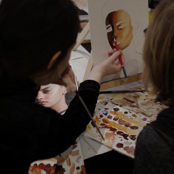Two people in black clothing face away from the camera as one of them uses a red pencil to draw in minor details in a half completed portrait. The table is littered with paint samples and reference images