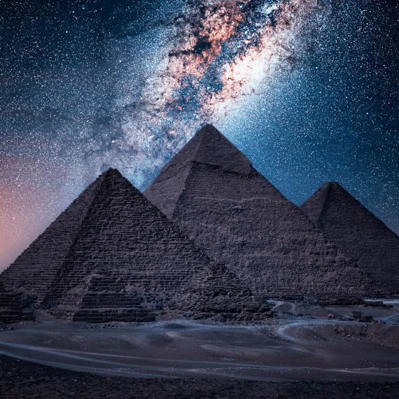 Three pyramids stand in darkness in a desert at night, backlit by the milkyway which shines vividly in the background in hues of orange, blue, purple and white, surrounded by hundreds of stars