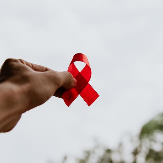 A hand holds a red ribbon folded over itself up against a bright sky. The red ribbon is symbolic for the World Aids Day awareness campaign.