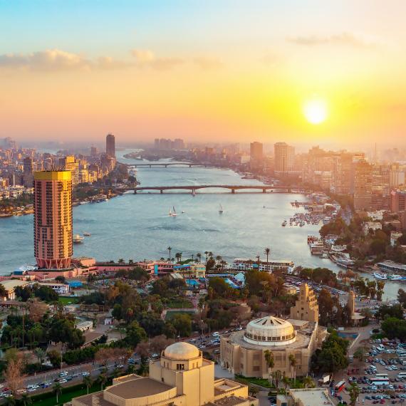 An idyllic panoramic view of the modern Cairo skyline, showing a sprawling city placed on either side of a large river. Under a bright yellow sunset, the towering skyscrapers and intricate temples appear to glow gold