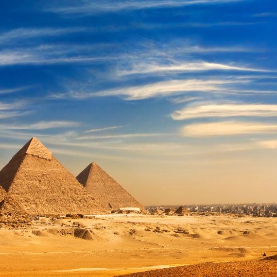 A vividly colourful landscape photograph of Giza, featuring the three large iconic pyramids, behind three smaller stone pyramids surrounded by golden yellow sandy desert plains and a bright blue sky