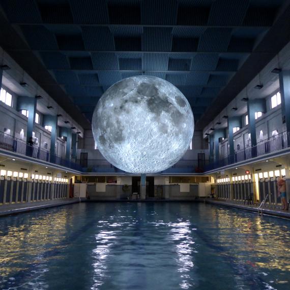 A large model of a moon glows with an etherial white light over a narrow public swimming pool which is backlit in white and yellow, creating a glowing reflection of colour in the otherwise darkened space
