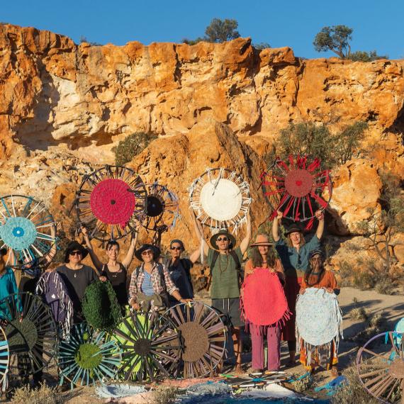 Small group of people dressed in outdoors clothing stand in front of a bright white and brown rocky hill and proudly display large circular weaving artworks in a variety of colours 