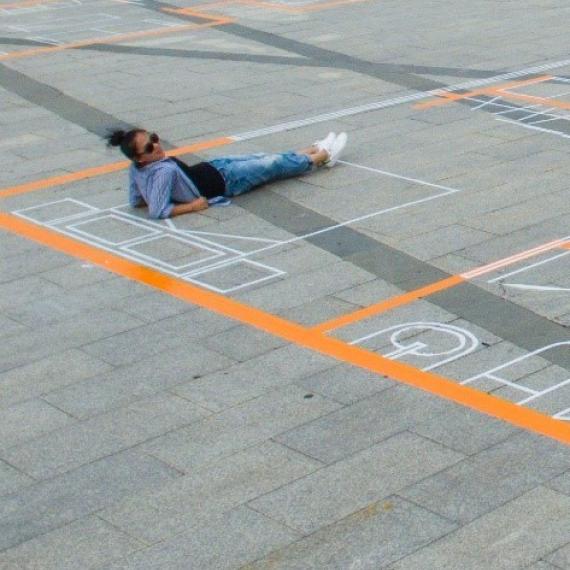 Different coloured tape is marked out on a concrete floor in thick orange and thin white. The tape is marking out the dimensions for a house. A woman in blue clothing in lying on markings for the bed and looking at he camera. 