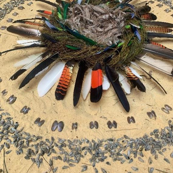 Circular artwork made from organic materials, including feathers, sand, plants and shells.  Centre piece of the work looks like a bird’s nest.  