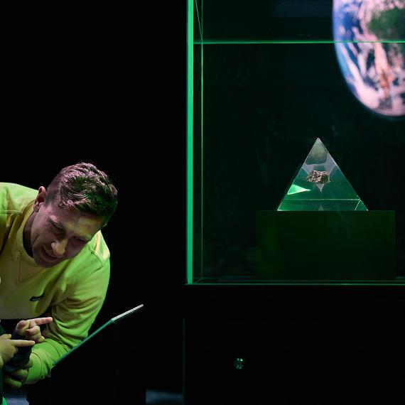 An adult and a young child with curly hair are bathed in green light from a Museum exhibition as they stare with interest at a showcase containing a glass pyramid holding a small section of moon rock.