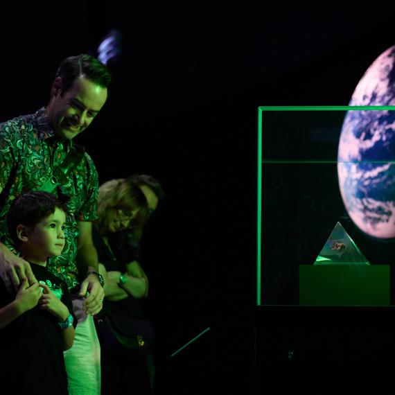 An adult and a young child are bathed in green light from a dramatically lit Museum exhibition. The adult smiles and looks down at the child as the child stares with interest at a glass pyramid containing a moon rock sample in a showcase.