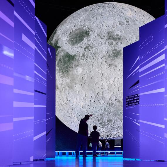 A large moon sculpture is visible down a corridor of brightly lit purple walls designed in a sci-fi aesthetic style with two people who appear small in comparison taking a selfie while in silhouette 