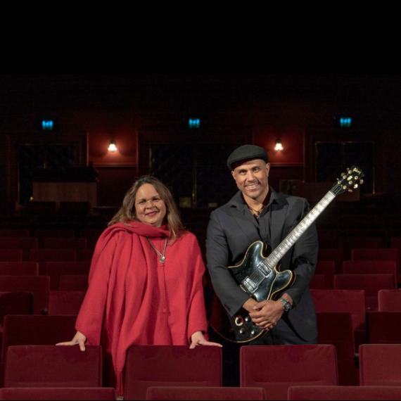 Musician's Gina Williams and Guy Ghouse stand among red velvet theatre chairs. Gina is wearing a red coat and silver necklace and Guy is wearing a black suit and hat, he is also holding a black guitar. Both are looking at the camera and smiling.  