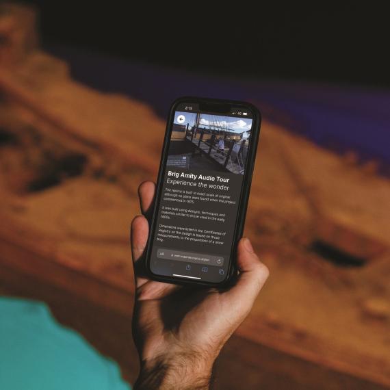 A hand holding a phone with the screen facing us. The screen shows the text Brig Amity audio tour with a picture of a walkway to walk to a ship. 