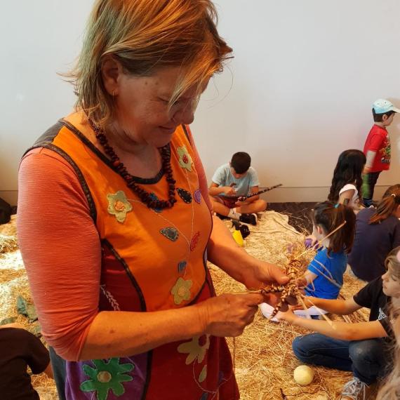 Woman standing weaving raffia with children sitting on floor behind her also weaving