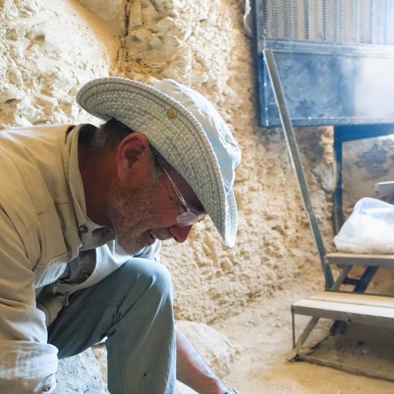 Man wearing hat and glasses crouched over ancient object brushing away dirt with paintbrush