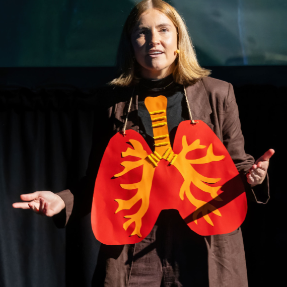 A blonde woman with cropped hair wears a paper heart around her neck, describing the anatomy 