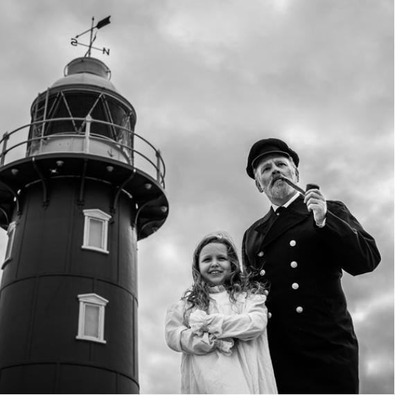 a black and white image of a lighthouse and two people standing in the foreground. One person is an adult, has a beard and wears a uniform smoking a pipe standing next to a young child with long hair and light coloured dress crossing their arms.