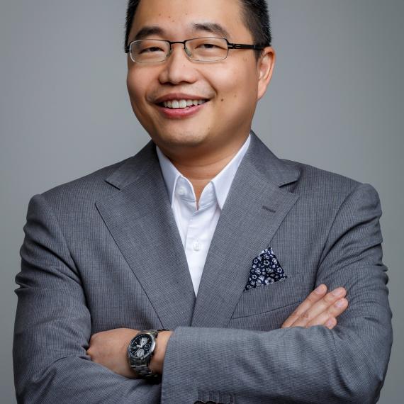 Marcus is an asian man with short black hair and glasses. He is wearing a grey suit and standing with his arms crossed smiling at the camera. 