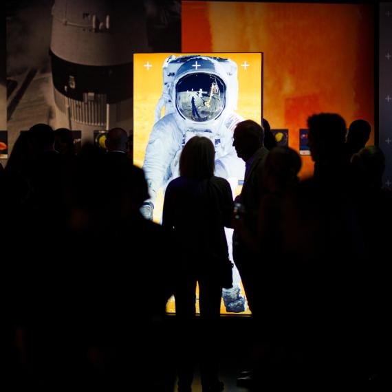 A brightly lit tall image of an astronaut in a darkened gallery surrounded by a large amount of silhouettes of people