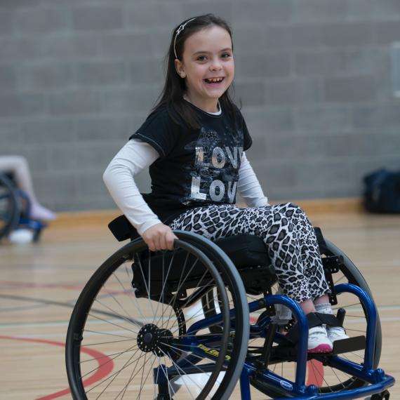 Girl in wheelchair and holding the wheels and smiling. Grey brick wall pictured in the background and a wooden floor with painted lines, indicating she is on a basketball court. 