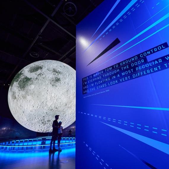 A dramatically lit large moon sculpture hangs in a dark gallery partially obstructed by a wall bathed in blue light with the lyrics to David Bowie's Space Oddity in black and white text