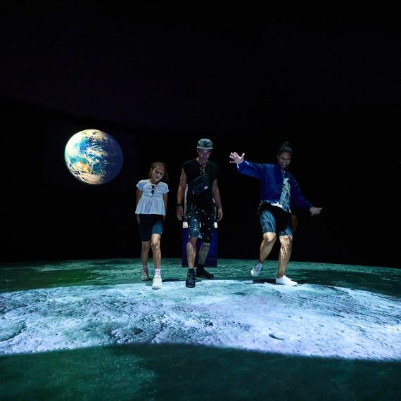 Three people walk over a projected image of the moon surface with footsteps. The installation sits in a darkened Museum gallery with a projection of the earth on the back wall.