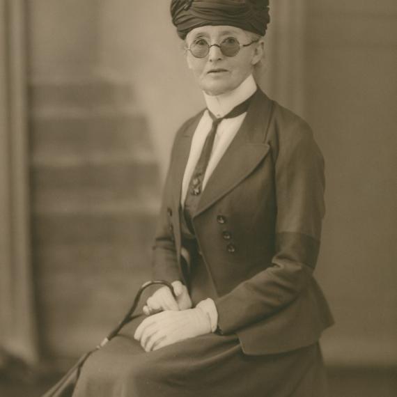 a woman dressed in posh clothing sits with hands crossed on her lap. The photograph is black and white.