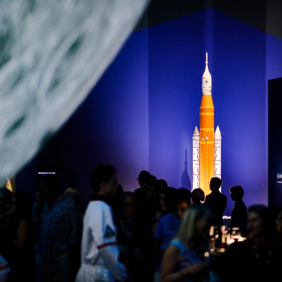 A orange and white model of a rocket is brightly lit in a darkened Museum space. A section of a large moon sculpture shines in the left hand side of the frame and silhouettes of people mill around beneath it.