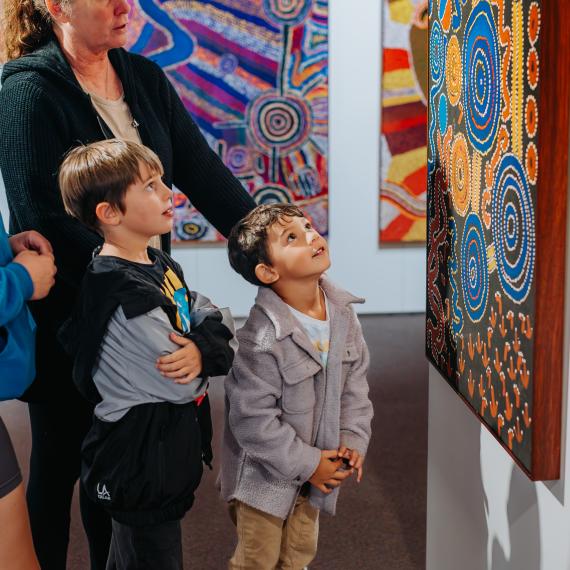 two oung children with their adults are looking at large artworks on display painted in traditional methods with vibrant colours 