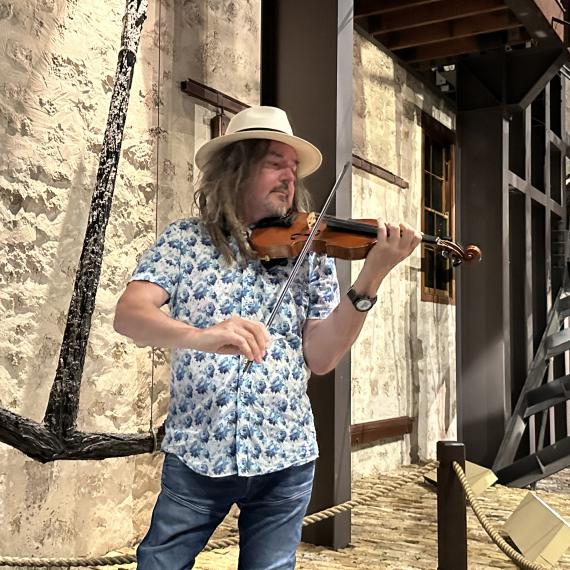An adult person with a hat stands with a violin in a position to play the instrument In the background is an anchor and part of a shipwreck