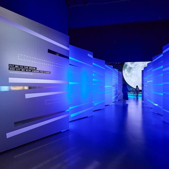 A hallway illuminated by coloured lights with reflective graphics leading to a large moon display.