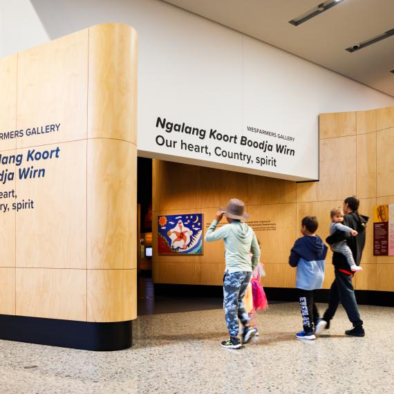 Entrance to Wesfarmers Gallery with wood paneling and text 'Ngalang Koort Boodja Wirn: Our heart, Country, spirit.' Five visitors approach the entrance.