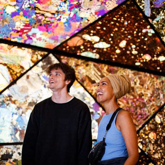 Two people smiling and looking upwards in a colorful, geometric, and illuminated space with intricate patterns.