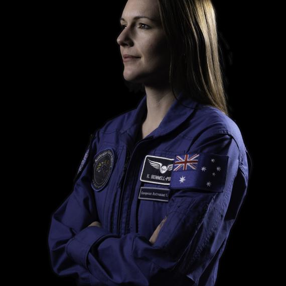 A woman in a blue astronaut suit with crossed arms, faces left.