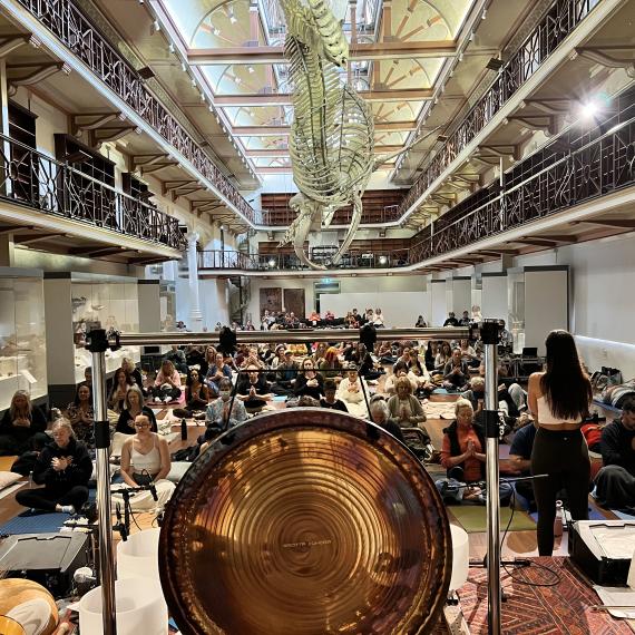 A large gong takes place on a stage surrounded by musicians. An immersed audience faces the stage among the balcony's, stairs and exhibits of Hackett Hall. 