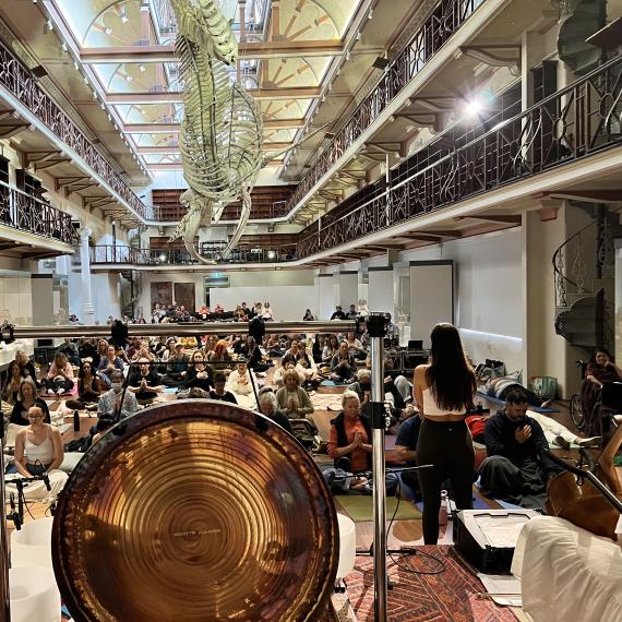 A large gong takes place on a stage surrounded by musicians. An immersed audience faces the stage among the balcony's, stairs and exhibits of Hackett Hall. 