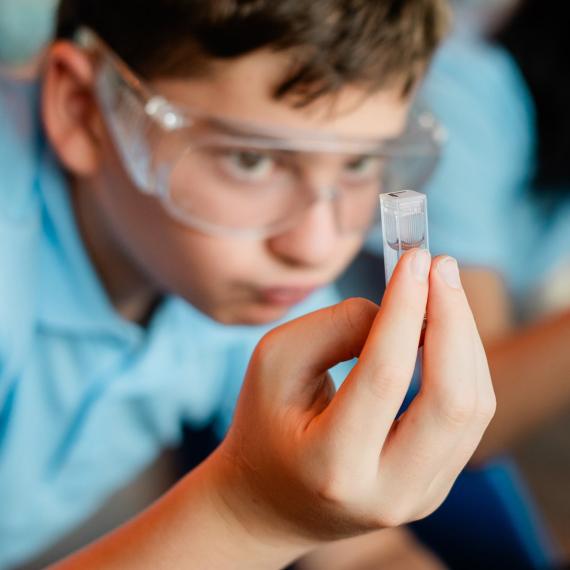 Boy wearing safety glasses inspects small scientific sample of marine life in a vial.