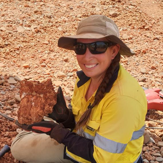 Lady wearing hat and sunglasses crouched, smiling at camera holding fossil