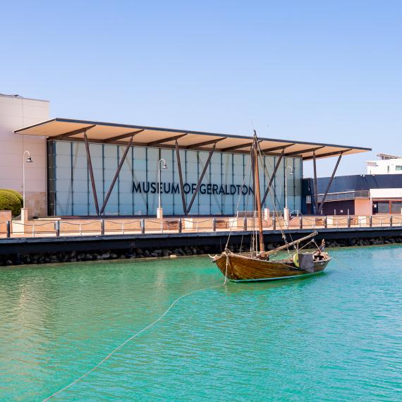 The facade of the Museum of Geraldton located on a harbour with vivid green-blue water and a vintage-style rowboat anchored at the front