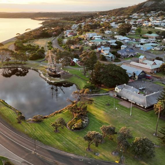 An aerial view of the Museum of the Great Southern site in Albany showing an expansive oceanic shore bordered by a grey road on the other side of which a large ships stands alongside a lake that reflects a black light in the slightly gloomy, grey day. A town is visible in the background