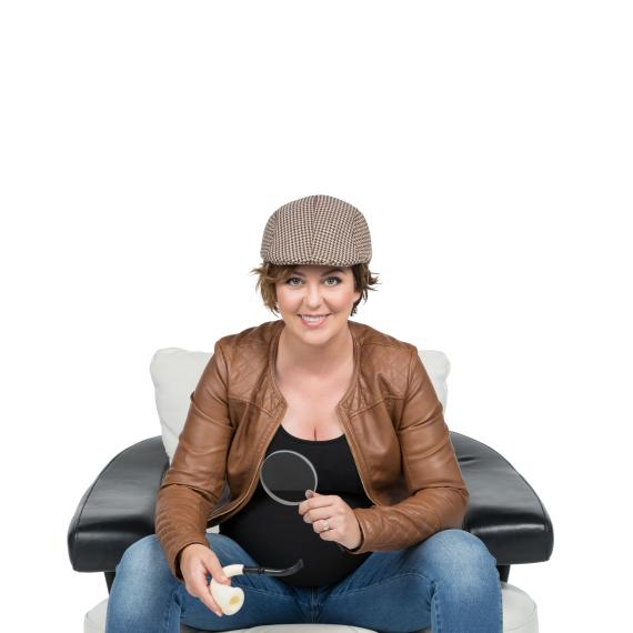 Paola is sitting in a black and white chairs facing the camera. She is smiling at the camera and wearing a Sherlock style hat while holding a pipe and magnifier glass.