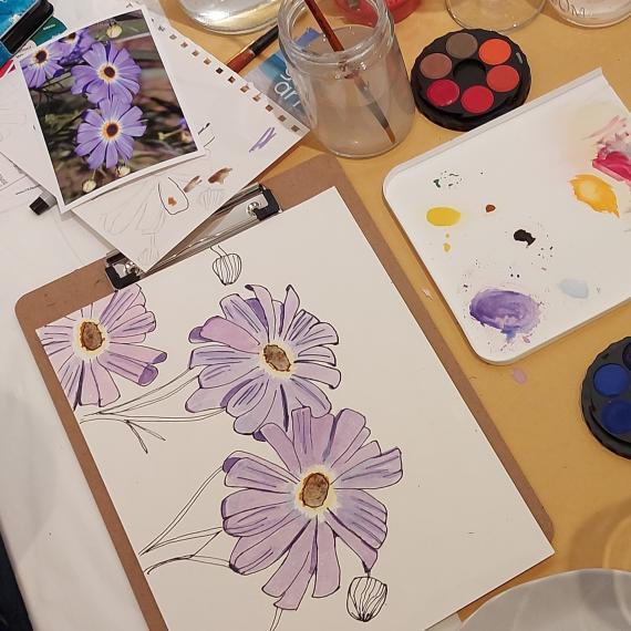 This image shows an example of a purple wildflower painted using watercolour paints. It also has some paints and a water jar with paintbrush next to it.