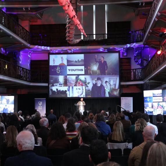 A large hall filled with people all facing a large screen and a male speaker at a lectern