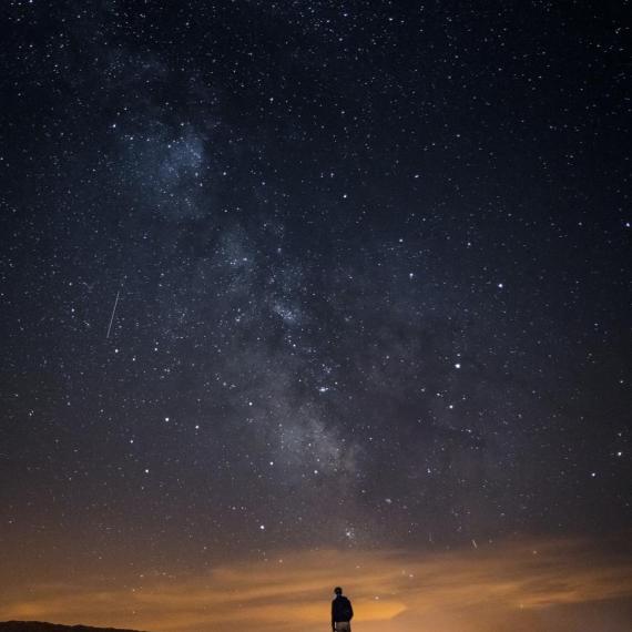 Image shows the night sky with many stars. The sun is in thwe final stages of settling on the horizon. A person is silhouetted in the distance. 