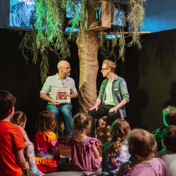 two adults on astage sit and read a story to a crowd of young children