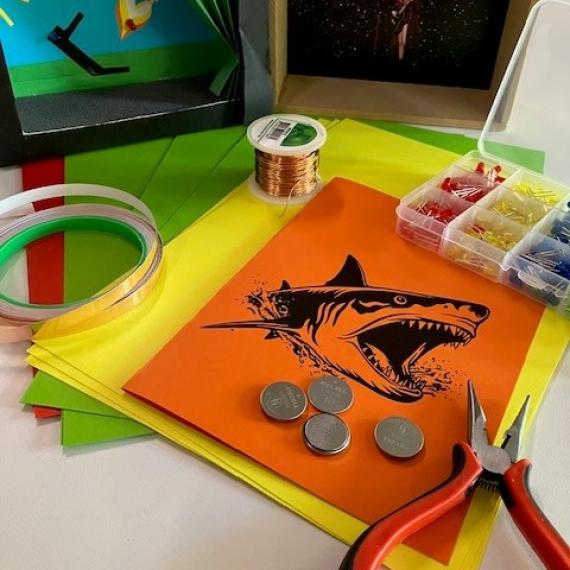 A range of materials are displayed on a table top. There is coloured card, LED lights, coin cell batteries , copper tape and some mini pliers. These will be used to create a small theatre using an electrical circuit to create lighting for the theatre.