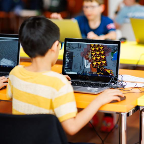 The image is looking over the right hand shoulder of  a young person using a laptop to play Minecraft