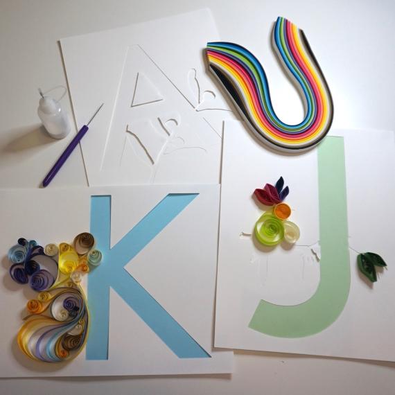 The image shows several items used in the craft of quilling. In particular strips of coloured paper, a quilling tool and the letter K and J letter templatescoloured s