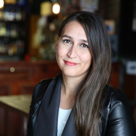 a person with long dark brown hair smiles at the camera wearing a white top and black jacket