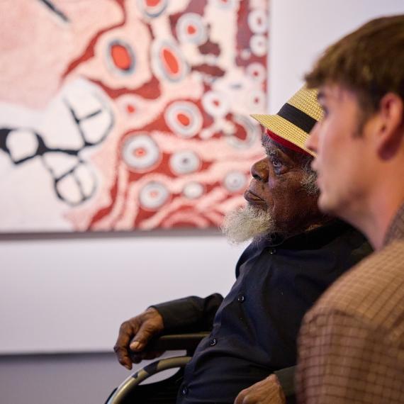 Old man in a wheelchair with an younger man next to him looking at a vibrant Aboriginal painting in an exhibition  