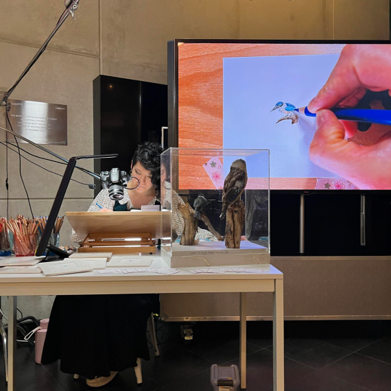 a person sitting at a desk drawing in detail with a screen behind showing the details 