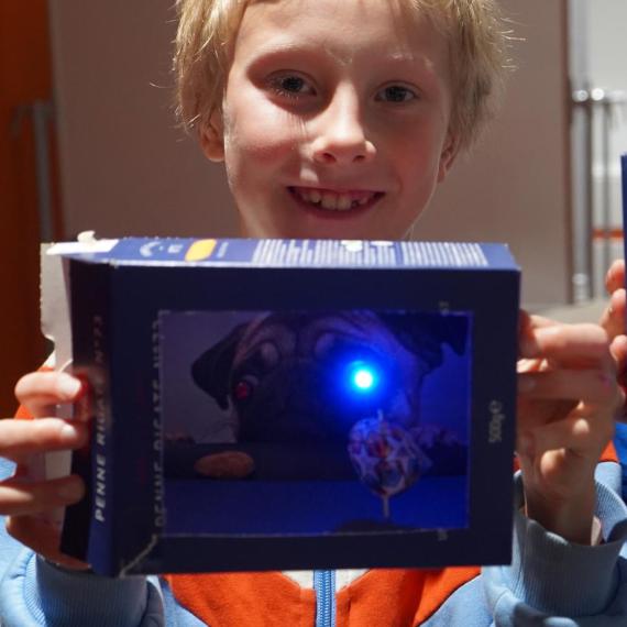 A young boy is holding up the theatre he made. It has blue LED lights in the display.