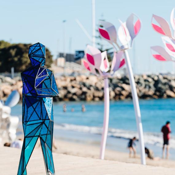 Blue glass artwork of a person standing on the beach in a thinking pose, with large white and pink flower structures in the distance. People can also be seen in the distance near the ocean. 